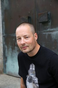 Tom Hillenbrand, der Autor: Man sieht einen Mann, ca. 30 Jahre alt. Er hat kurz rasierte Haare und einen Dreitage-Bart. Er hat Ohrringe. Er lächelt in die Kamera. Im Hintergund sieht man ein große Tür. Er hat ein dunkles T-Shirt an.
