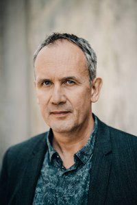 Volker Kutscher, der Autor: Ein Mann mittleren Alters mit grauen und schütterem Haar steht vor einer Mauer. Er hat ein dunkelblaues Sakko und ein gemustertes, blaues Hemd an. Er lächelt leicht in die Kamera.