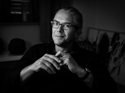 Ein Mann, Steffen Kopetzky, mit grauen Haaren und Brille sitzt leicht lächelnd und hält sich die eine Hand mit der anderen Hand.