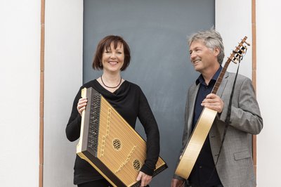 Eine Frau hält ein Hackbrett in den Händen und ein Mann hält eine Gitarre. Beide stehen nebeinander und lachen. Beide Personen tragen Abendkleidung.