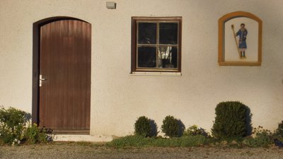 Hauseingang mit Fenster und einer Reliefstatue. Davor Kies und Büsche