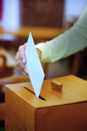 Eine Wahlurne in die eine Hand ein Kuvert wirft