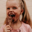 Mädchen mit blonden Zöpfen leckt ein Schokoeis. Das Eis tropft und ist über all in ihrem Gesicht und auf dem T-Shirt.