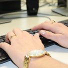 Zwei Hände tippen auf eine Tastatur. Die Person, dessen Hände auf die Tastatur tippen, tragen goldene Ringe, Nagellack und eine Uhr.
