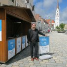 Ein Mann steht am Hauptplatz vor der Vorverkaufshütte und posiert mit einem Schild, aufdem "Jetzt Ticket sichern" steht. Das Logo ist blau.