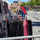 Absperrungen befinden sich um einige Stände mit Kleidung auf dem Hauptplatz. Ein Mann mit Maske schaut sich in der Ferne um. Im Hintergrund ist das Rathaus zu erkennnen. Im Vordergrund sind Absperrungen und verschiedenste Hosen auf einer Kleiderstange. Auch ein Sakko hängt am Stand mit Anziehsachen.