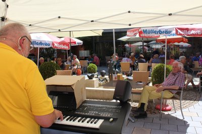 Mann mit gelben Shirt spielt auf einem schwarzen Keyboard. Vor ihm sitzen viele Rentner auf einem Platz mit Schirmen. In der Mitte dieses Platzes befindet sich ein Brunnen mit verschiedenen Figuren.