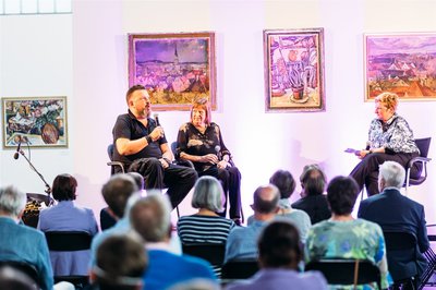 Drei Menschen sitzen vor einem Publikum und vor Gemälde von Weingartner auf einer Bühne. Zwei davon sind Familienmitglieder der Familie Weingartner und die Dritte interviewt die Mitglieder.