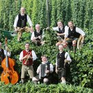 Die Dellnhauser Musikanten zwischen Hopfen. Sie tragen alle Tracht und jeder hält ein Instrument in der Hand. Darunter sind ein Kontrabass, eine Trompete und eine Klarinette. Alle außer einer haben eine schwarze Weste an.