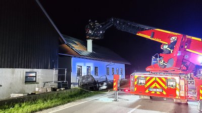 Ein Drehleiterfahrzeug der Feuerwehr steht nachts vor einem eingeschossigen Haus. Im Vordergrund am Haus ein ausgebranntes Auto