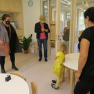 In einem Kindergartenraum  stehen vier Erwachsene und ein Kind. Das kleine Mädchen in einem gelben Kleid steht an einem Tisch gelehnt. Auf dem Bild sind Katrin Maier, Roland Dörfler Simona Schmutterer, das Mödchen und eine Erzieherin zu sehen.