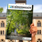 Hand, die das Bürgermagazin in die Höhe hält. Dahinter das Rathaus