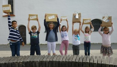 Sieben Kinder, ca. zwischen 6 und 8 Jahren, halten Pakete mit den Gewinnen aus dem Lesewettbewerb in die Höhe. 3 Buben und 4 Mädchen sieht man. Alle Kinder lächeln..