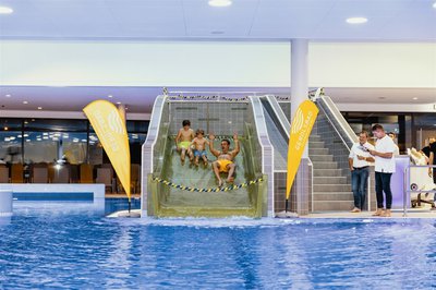 Ein Hallenbad. Auf der Rutsche ein Mann in gelber Badehose und zwei Kinder
