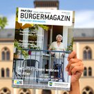 Hand hält das aktuelle Bürgermagazin zum Thema Selbstbestimmt wohnen im Alter in die Höhe. Auf dem Titelbild ist eine ältere Dame auf einem Balkon.