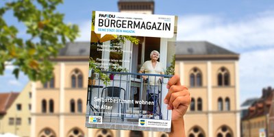 Hand hält das aktuelle Bürgermagazin zum Thema Selbstbestimmt wohnen im Alter in die Höhe. Auf dem Titelbild ist eine ältere Dame auf einem Balkon.
