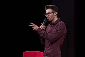 Mann in dunkelroten Hemd auf einer Bühne mit einem Mikro in der Hand.