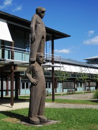 Zwei Figuren übereinander vor einem Gebäude im Gras