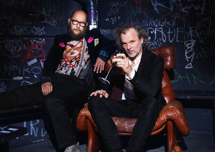 Zwei Männer sitzen auf einem alten Ledersessel und im Hintergrund ist eine Wand mit Graffiti.