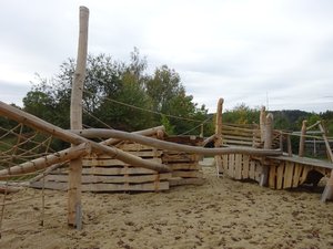 Spielplatz mit Holz-Spielgeräten und Sand, im Hintergrund Bäume und Hecken