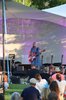 Musikerin Claudia Koreck mit blauem Sommerkleid und blondem schulterlangen Haar. Sie singt in ein Mikrofon und spielt auf einer roten E-Gitarre. Zuschauer sitzen vor der Bühne bei diesem Kultursommer Picknick-Konzert im Pfaffenhofener Bürgerpark.