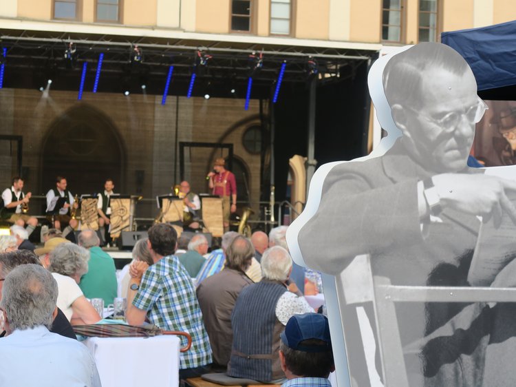Menschen an Bierbänken auf dem Hauptplatz; im Hintergrund eine Bühne und rechts ein Aufsteller von Joseph Maria Lutz.
