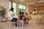 Blick in die Küche, Kinder sitzen mit einer Erzieherin an einem runden Holztisch und essen.