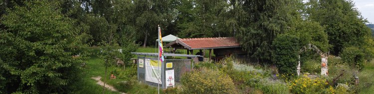 Bäume, dazwischen eine Gartenlaube mit einer fahne und Informationsschildern zum Interkulturgarten. REchts im Bild ein bepflanzter Torbogen