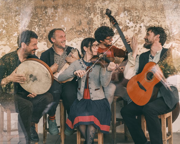 Band sitzt vor einer Wand auf Stühlen und hat Instrumente in der Hand.