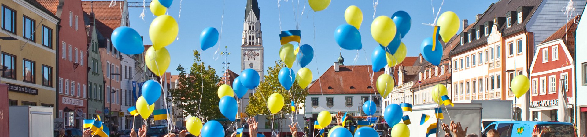 Pfaffenhofener Hauptplatz im sonnenschein mit bunen Häusern, Kirche im Hintergrund, gelbe und blaue Luftballons steigen auf