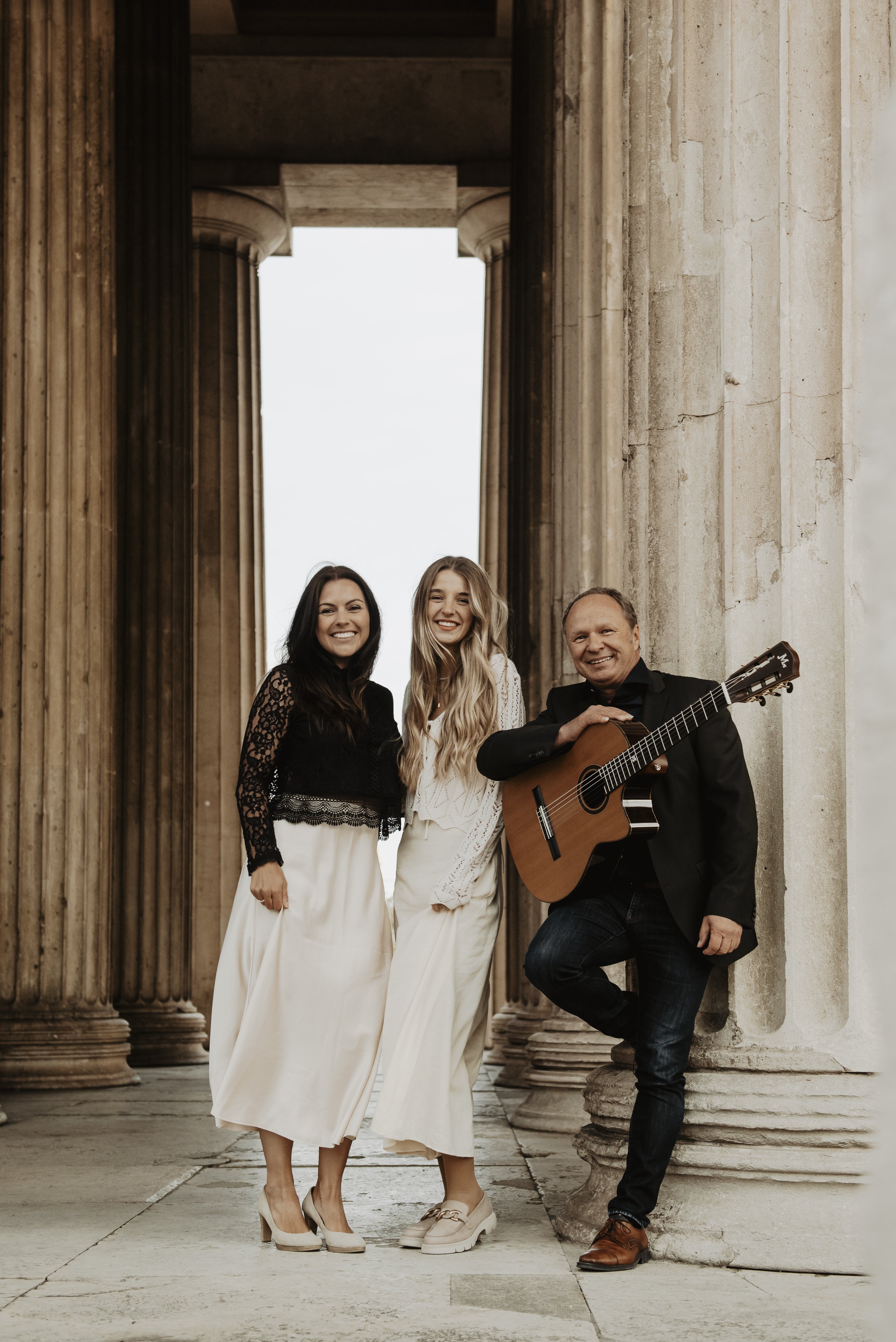 Zwei Frauen und ein Mann mit Gitarre stehen an einer weißen Säule.
