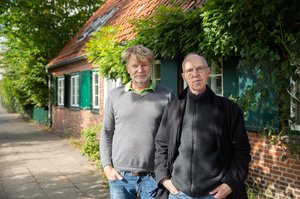 Die beiden Autoren vor einem Backstein-Haus mit grünen Fensterläden.