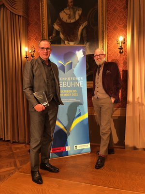 Auf der Pfaffenhofener Lesebühne. Der Autor Volker Kutscher und der Moderator des Abends Thomas Boehm stehen im Festsaal vor einem Rollup. Volker Kutscher hat sein Buch in der Hand.