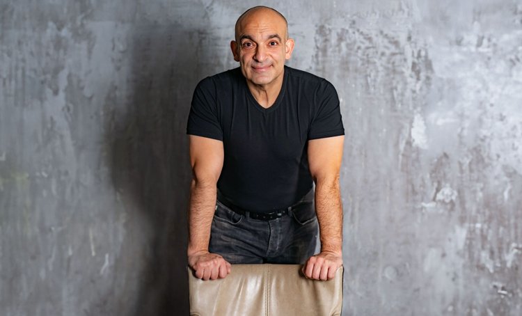 Django Asül vor einer grauen Steinwand, trägt ein schwarzes T-Shirt und stützt sich auf einem Stuhl ab
