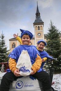 ZWei Gelb-blau gekleidete Weihnachtswichtel mit einem Sack vor dem Rathaus. Dahinter Christbäume