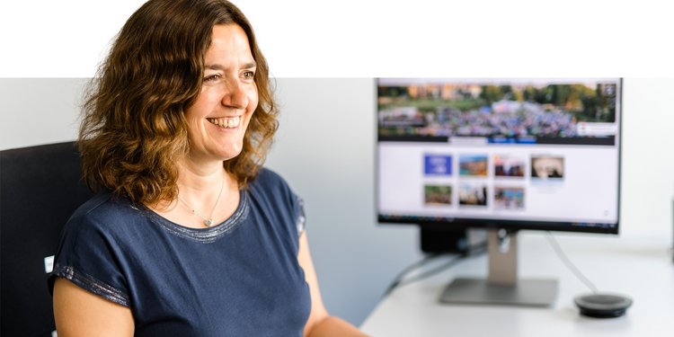 Eine Frau in blauem Shirt sitzt am Arbeitsplatz; im Hintrgrund ein PC-Bildschirm
