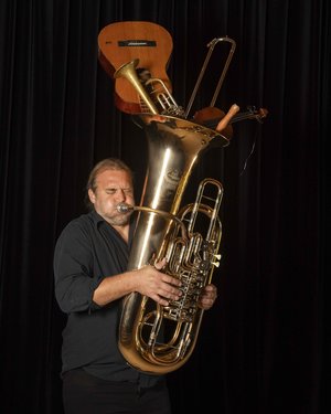 Hofmeir spielt die Tuba, oben in der Tuba stecken andere Instrumente wie eine Gitarre, Hintergrund ist schwarz