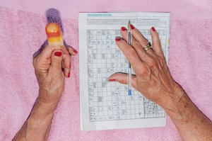 Hände einer älteren Dame auf pinkem Handtuch mit Eis und Kreuzworträtsel